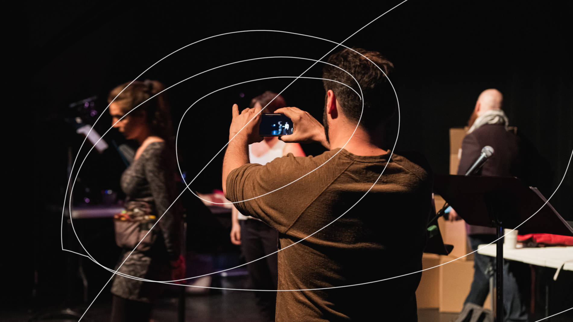 Spectacle "Zombies Westerns de la terreur": En avant-plan, un homme tient un appareil photo et filme une femme marchant vers l'avant de la scène. À la droite de cet homme se trouvent un micro et un lutrin. En arrière-plan, deux acteurs sont en mouvement.