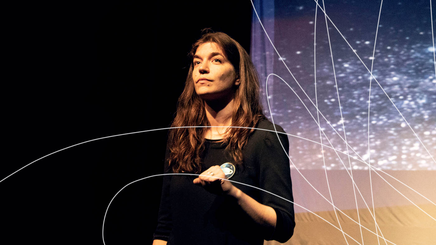 Une actrice aux cheveux long et brun vêtue d'un gilet noir orné d'un écusson avec une image de planète, debout sur la scène, regarde au loin. Superposé à l'image, un trait artistique qui rappelle le logo de Théâtre Déchaînés.