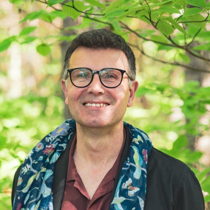 Portrait d'un homme souriant, lunette noir, yeux bleus et cheveux foncés.