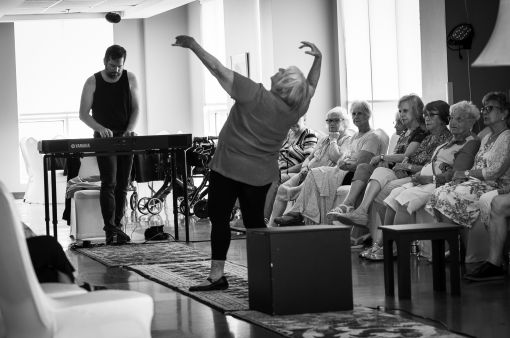 Un comédien joue du piano et une comédienne fait de grands mouvements de danse devant lui et le public.