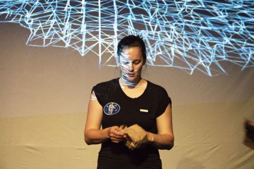 Andrée-Anne tient un sac de papier dans ses mains, des lignes bleus se dessinent sur l'écran derrière elle.